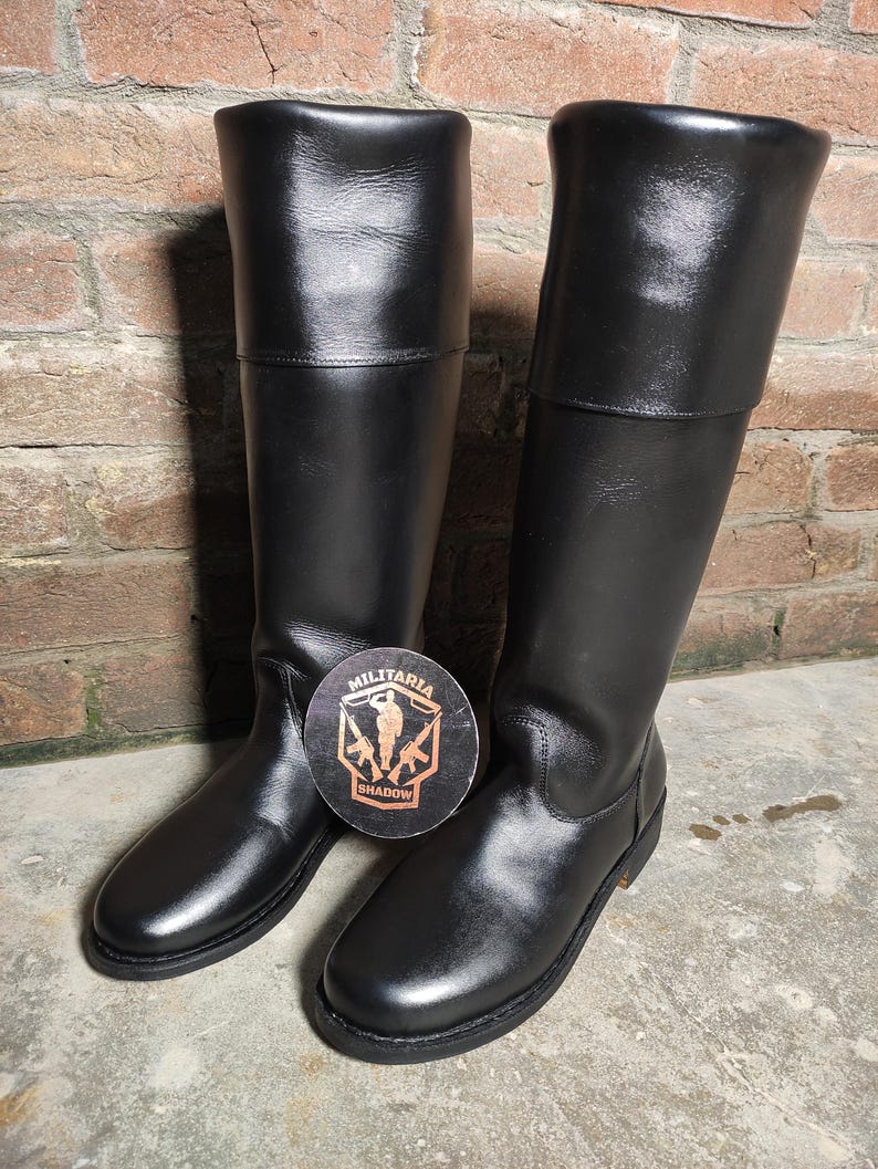 May include: A pair of black leather boots with a tall shaft and a folded-over top. The boots are shiny and appear to be new. A round sticker with the words "MILITARIA SHADOW" and a military-themed graphic is placed between the boots.