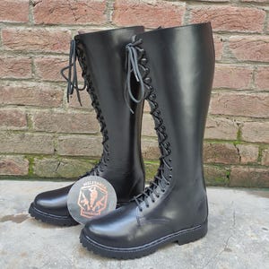 May include: A pair of tall, black leather boots with a lace-up design. The boots reach the knee and have a round toe and a thick sole. The boots are displayed in front of a brick wall. A logo is visible on the ground.