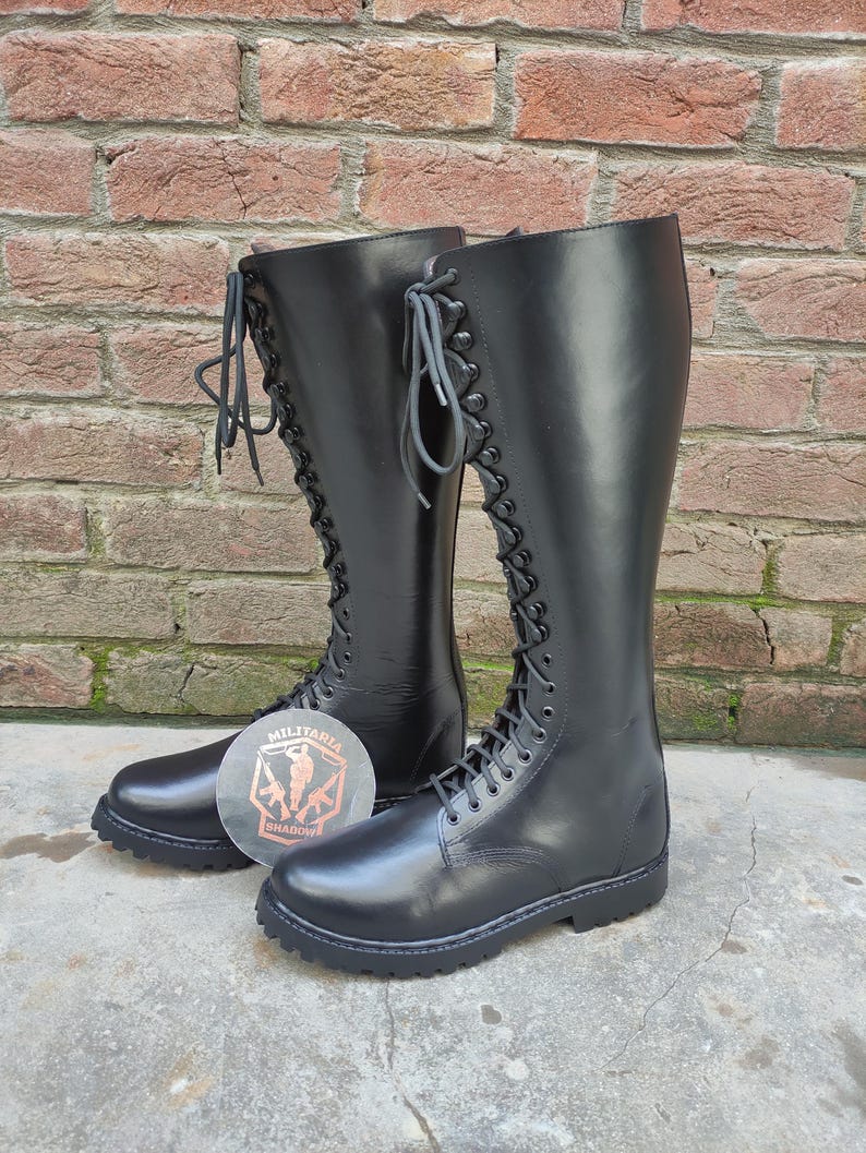 May include: A pair of tall, black leather boots with a lace-up design. The boots reach the knee and have a round toe and a thick sole. The boots are displayed in front of a brick wall. A logo is visible on the ground.