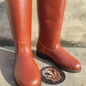 May include: A pair of tall, reddish-brown leather boots. The boots have a smooth finish, a rounded toe, and a low heel. A round emblem with the text "MILITARIA SHADOW" is visible near the boots.