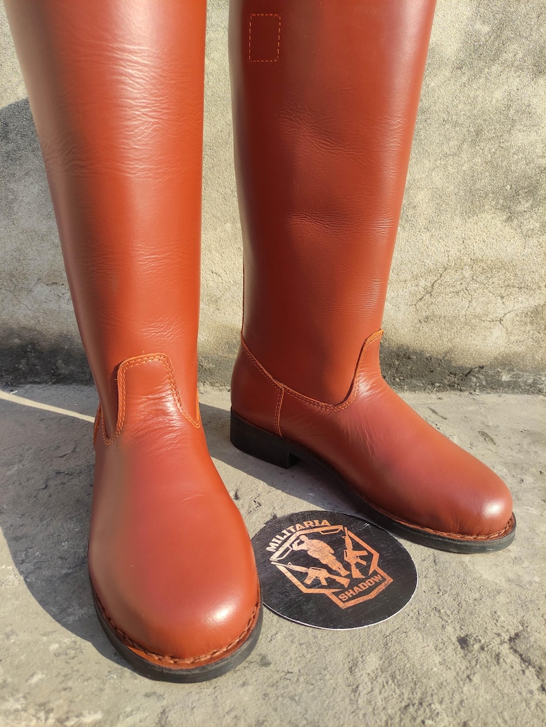 May include: A pair of tall, reddish-brown leather boots. The boots have a smooth finish, a rounded toe, and a low heel. A round emblem with the text "MILITARIA SHADOW" is visible near the boots.