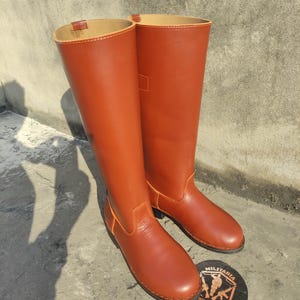 May include: A pair of tall, reddish-brown leather boots with a smooth finish. The boots have a classic design, with a rounded toe and a high shaft. Orange stitching accents the seams. A round emblem with the text "MILITARIA SHADOW" is visible.