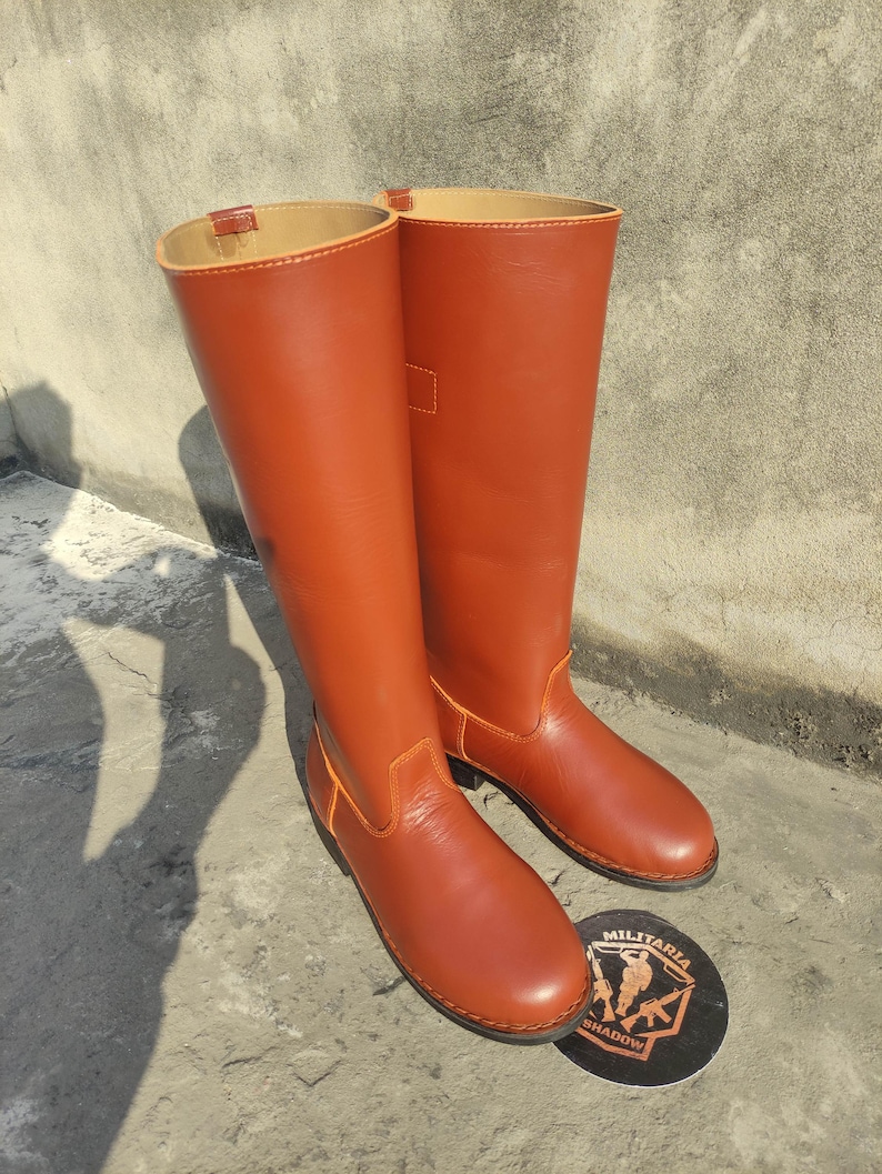 May include: A pair of tall, reddish-brown leather boots with a smooth finish. The boots have a classic design, with a rounded toe and a high shaft. Orange stitching accents the seams. A round emblem with the text "MILITARIA SHADOW" is visible.