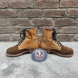May include: A pair of brown and tan suede boots with black laces. The boots have a two-tone design, with a darker brown on the toe and heel and a lighter tan on the upper. A round emblem with the text "MILITARIA SHADOW" is between the boots.