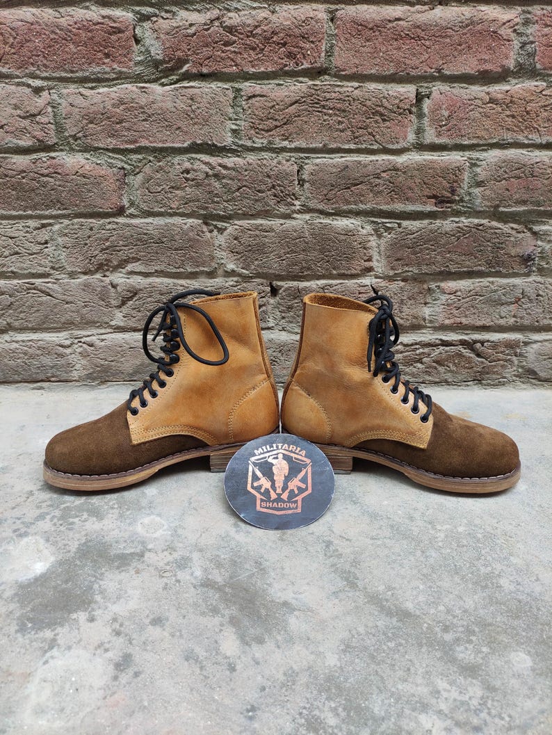May include: A pair of brown and tan suede boots with black laces. The boots have a two-tone design, with a darker brown on the toe and heel and a lighter tan on the upper. A round emblem with the text "MILITARIA SHADOW" is between the boots.
