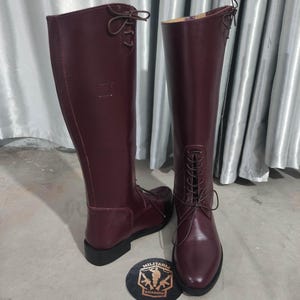 May include: A pair of tall, burgundy leather boots with laces and a rounded toe. The boots have a high shaft and a small heel. A round emblem with the text "MILITARIA SHADOW" is at the base of the boots.