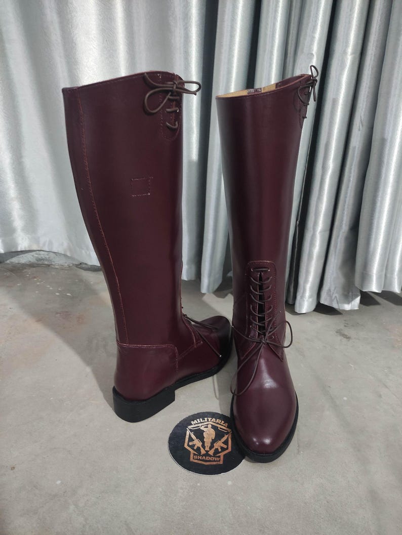 May include: A pair of tall, burgundy leather boots with laces and a rounded toe. The boots have a high shaft and a small heel. A round emblem with the text "MILITARIA SHADOW" is at the base of the boots.