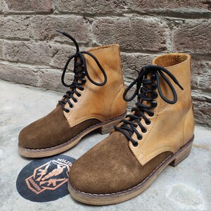 May include: A pair of two-tone ankle boots. The boots feature a tan leather upper with a brown suede toe and heel. Black laces thread through metal eyelets. The boots are set on a light brown sole.
