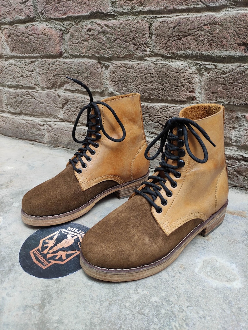 May include: A pair of two-tone ankle boots. The boots feature a tan leather upper with a brown suede toe and heel. Black laces thread through metal eyelets. The boots are set on a light brown sole.