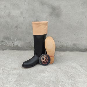 May include: Black leather riding boot with a tan leather cuff. A separate sole and a round emblem with text and a military design are also present. The boot is tall and appears to be new.