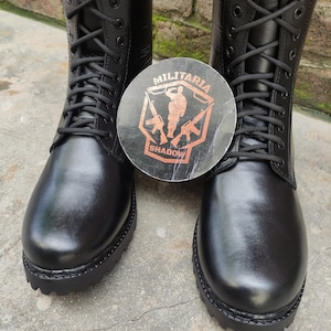 May include: Pair of black leather combat boots with a lace-up design. The boots have a thick sole and are displayed with a round sticker featuring the text "MILITARIA SHADOW" and a military-themed graphic.