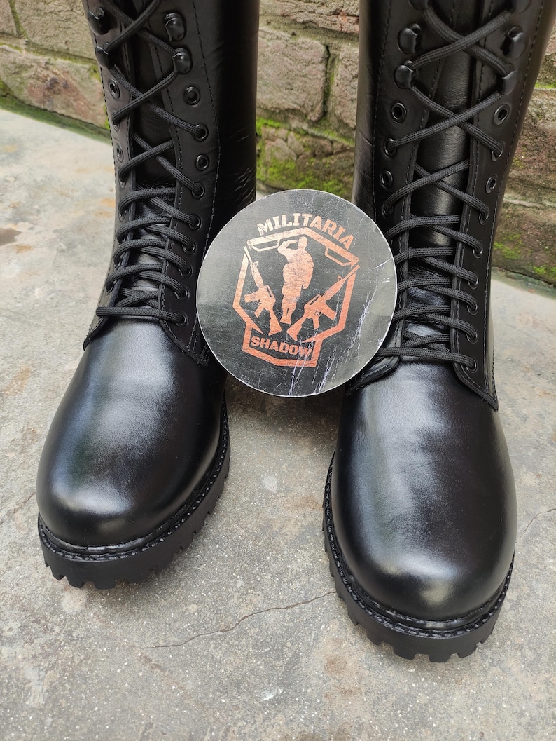 May include: Pair of black leather combat boots with a lace-up design. The boots have a thick sole and are displayed with a round sticker featuring the text "MILITARIA SHADOW" and a military-themed graphic.