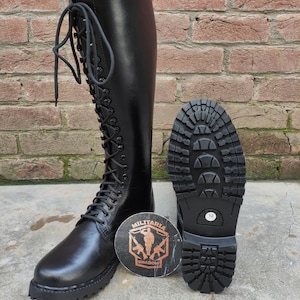 May include: Black knee-high combat boot with a lace-up design. The boot is displayed next to its sole, showcasing a tread pattern. A round emblem with the text "MILITARIA SHADOW" is also present.