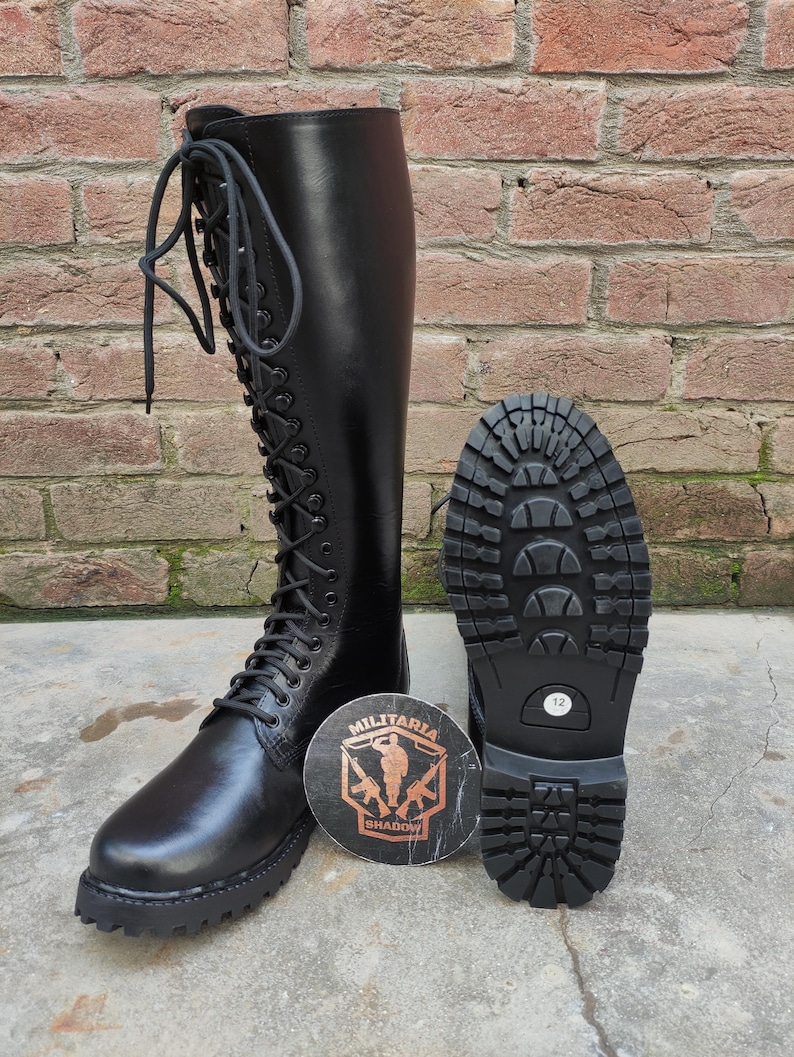 May include: Black knee-high combat boot with a lace-up design. The boot is displayed next to its sole, showcasing a tread pattern. A round emblem with the text "MILITARIA SHADOW" is also present.