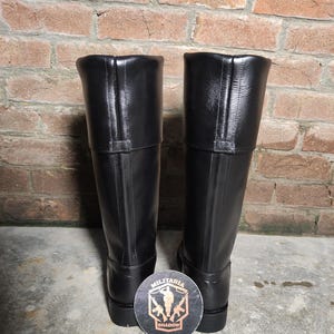 May include: A pair of tall, black leather boots with a smooth finish. The boots have a knee-high design with a folded-over top and a small strap detail. A round coaster with the text "Militaria Shadow" is at the base of the boots.