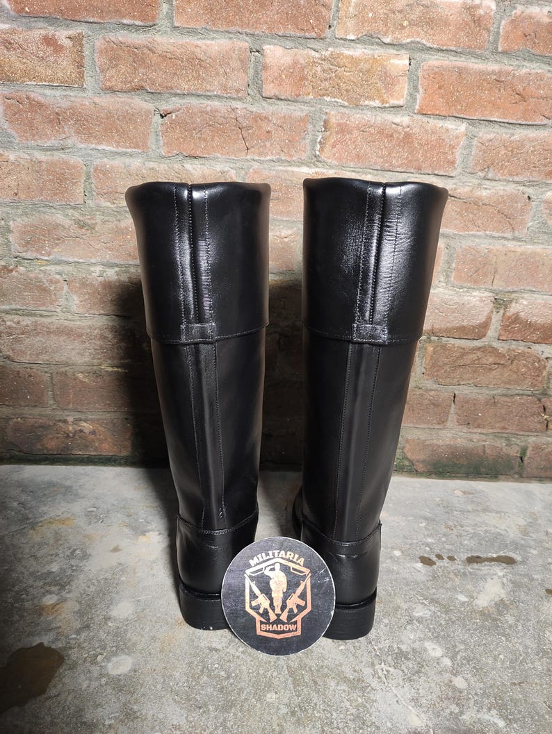 May include: A pair of tall, black leather boots with a smooth finish. The boots have a knee-high design with a folded-over top and a small strap detail. A round coaster with the text "Militaria Shadow" is at the base of the boots.