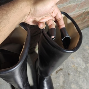 May include: A pair of black leather boots with a smooth, shiny finish. The boots are tall, reaching up to the mid-calf, and have a classic design. The interior lining is a light beige color. A sticker with the word "MILITARIA" is visible.