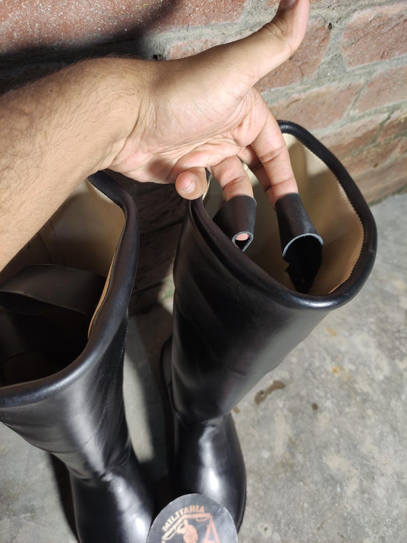 May include: A pair of black leather boots with a smooth, shiny finish. The boots are tall, reaching up to the mid-calf, and have a classic design. The interior lining is a light beige color. A sticker with the word "MILITARIA" is visible.