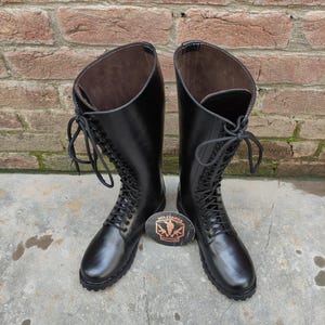 May include: A pair of tall, black leather boots with lace-up fronts. The boots have a smooth, polished finish and are set against a brick wall background. A small, oval emblem with text is placed between the boots.
