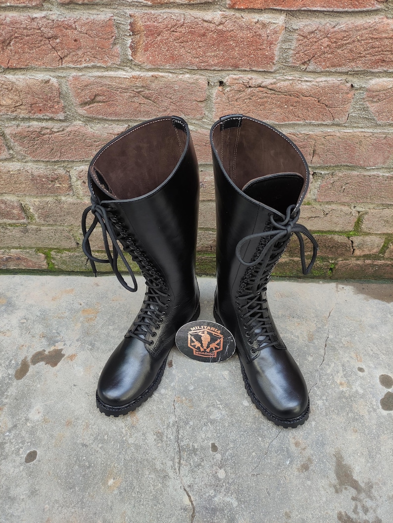 May include: A pair of tall, black leather boots with lace-up fronts. The boots have a smooth, polished finish and are set against a brick wall background. A small, oval emblem with text is placed between the boots.