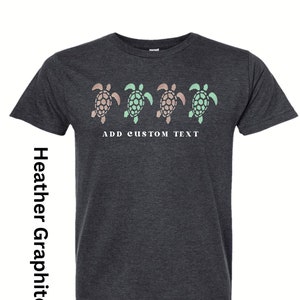 May include: A dark heather grey t-shirt featuring a row of three sea turtle silhouettes in shades of green and pink. The text "ADD CUSTOM TEXT" is printed below the turtles.