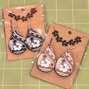 May include: Two pairs of teardrop-shaped earrings. One pair is black and white, the other is brown and white. Each earring features a white hibiscus flower and tribal patterns. The earrings are displayed on brown cards with floral accents.