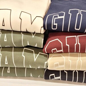 Op de afbeelding: Stapel van vijf T-shirts met het woord "GUAM" in wit op elk shirt. De shirts zijn in verschillende kleuren: wit, groen, beige, bordeauxrood en donkerblauw.