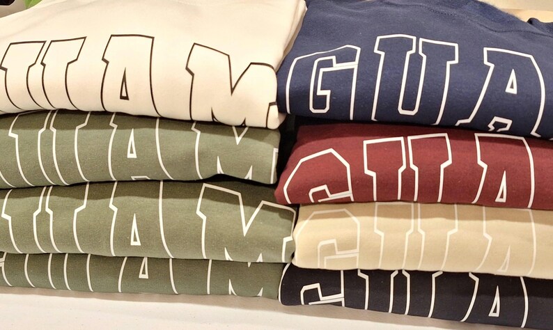 Op de afbeelding: Stapel van vijf T-shirts met het woord "GUAM" in wit op elk shirt. De shirts zijn in verschillende kleuren: wit, groen, beige, bordeauxrood en donkerblauw.