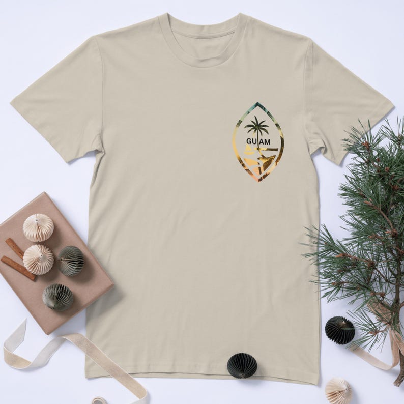 Pu&ograve; includere: Una t-shirt beige con una grafica della bandiera di Guam. La bandiera &egrave; una foglia blu e verde con una palma e la parola "GUAM" in testo nero.