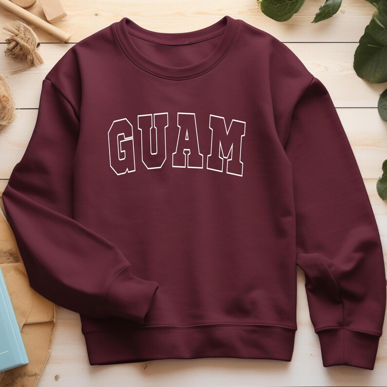 Op de afbeelding: Bordeauxkleurige trui met witte tekst die "GUAM" zegt.