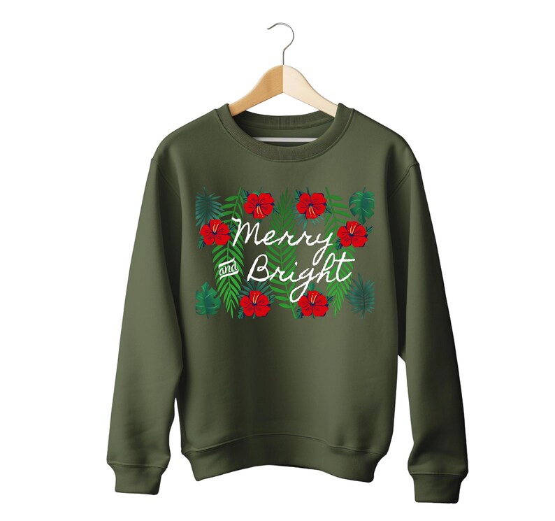 May include: An olive green sweatshirt with the words "Merry and Bright" in white script, surrounded by red hibiscus flowers and green leaves. The sweatshirt is hanging on a wooden hanger.