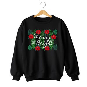 May include: Black sweatshirt with a festive design. The design features red hibiscus flowers and green leaves surrounding the words "Merry and Bright" in white script. The sweatshirt is hanging on a wooden hanger.