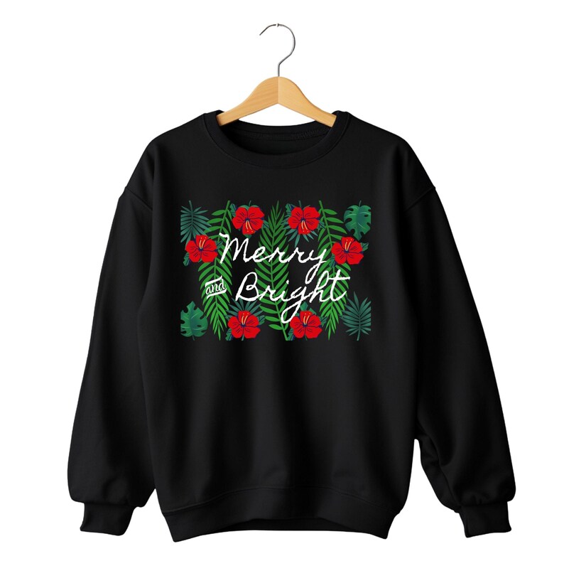 May include: Black sweatshirt with a festive design. The design features red hibiscus flowers and green leaves surrounding the words "Merry and Bright" in white script. The sweatshirt is hanging on a wooden hanger.