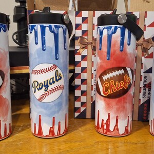 Op de afbeelding: Vier roestvrijstalen bekers met verschillende sportteamontwerpen. De bekers zijn wit met blauw en rood waterverfachtige ontwerpen. De ontwerpen omvatten een honkbal met "Royals" erop geschreven, een voetbal met "Chiefs" erop geschreven en een honkbal met "Royals" erop geschreven. De bekers hebben zwarte deksels met een handvat.