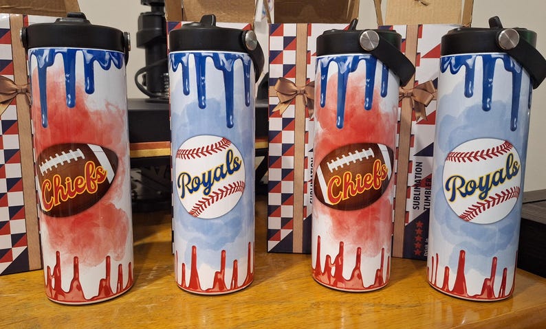 Op de afbeelding: Vier roestvrijstalen bekers met verschillende sportteamontwerpen. De bekers zijn wit met blauw en rood waterverfachtige ontwerpen. De ontwerpen omvatten een honkbal met "Royals" erop geschreven, een voetbal met "Chiefs" erop geschreven en een honkbal met "Royals" erop geschreven. De bekers hebben zwarte deksels met een handvat.