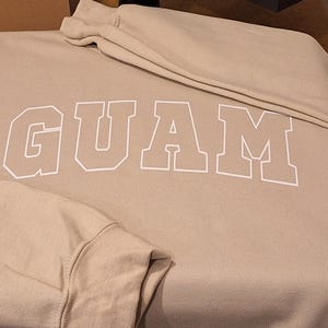 Op de afbeelding: Een beige sweatshirt met het woord "GUAM" in wit op de voorkant.