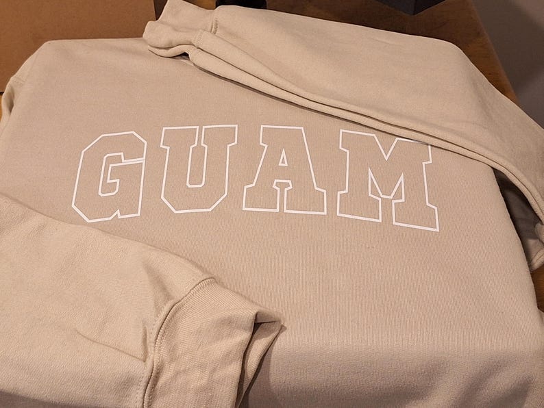 Op de afbeelding: Een beige sweatshirt met het woord "GUAM" in wit op de voorkant.