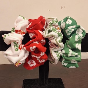 May include: Four fabric scrunchies in a variety of floral patterns. The scrunchies are in white, red, and green, with floral designs. The scrunchies are displayed on a black stand. The scrunchies are a hair accessory.