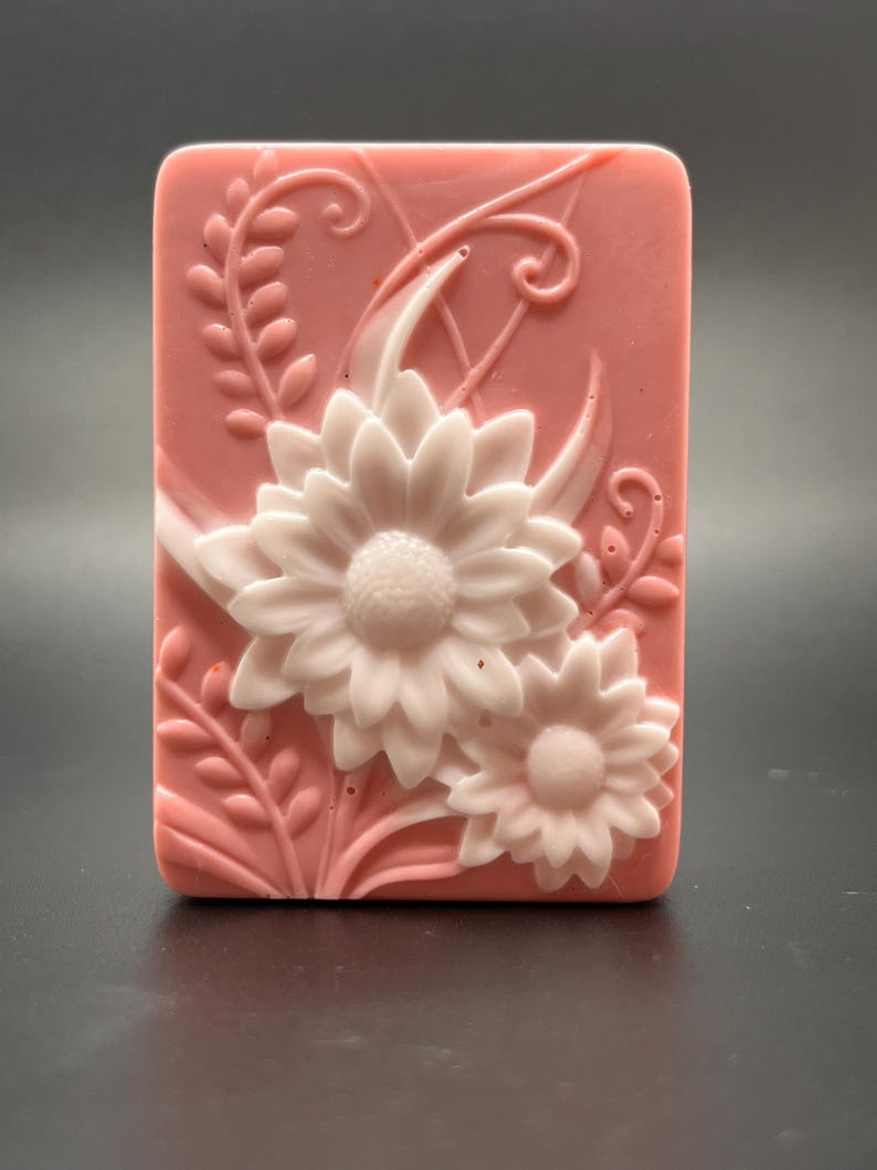 May include: A pink soap bar with white floral designs. The soap bar has a raised design of two flowers with leaves and swirls.