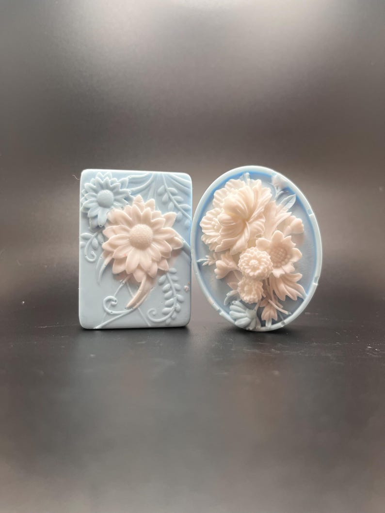 May include: Two decorative soap bars with floral designs. The soap bars are light blue with white raised floral designs. One soap bar is rectangular and the other is oval.