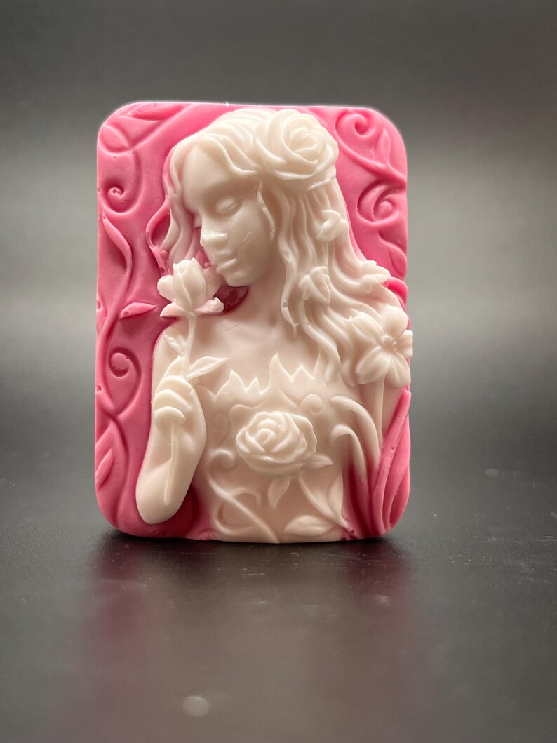 May include: A white soap bar with a detailed carving of a woman with long hair and a flower in her hand. The soap is set against a pink background with floral designs.