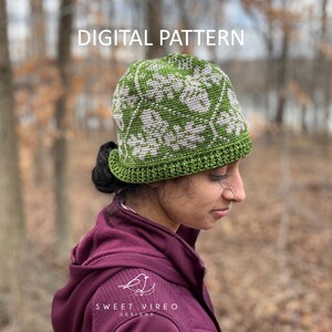 May include: A green and white crocheted hat with a floral design. The hat is worn by a person in a maroon jacket. The words "DIGITAL PATTERN" are at the top of the image. The Sweet Vireo Designs logo is on the jacket.