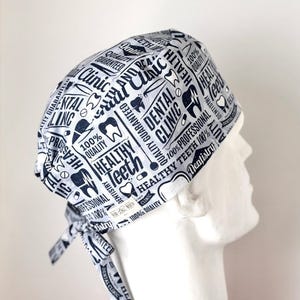 May include: A light gray and white dental scrub cap with a repeating pattern of dental-themed text and graphics. The text includes phrases like "Dental Clinic," "Healthy Teeth," and "100% Quality." The cap is tied at the back.