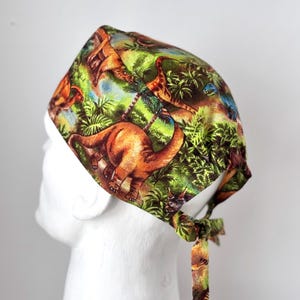 May include: A green, brown, and blue patterned scrub hat with a dinosaur print. The hat has a tie at the back.