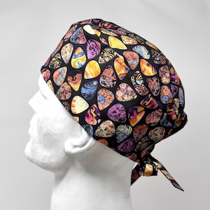 May include: A black surgical scrub cap featuring a colorful pattern of abstract shapes in shades of orange, yellow, purple, and blue. The cap is tied at the back and is designed to cover the head. The fabric has a repeating design.
