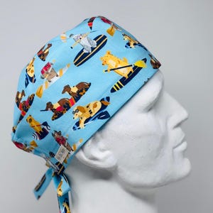 May include: A light blue scrub cap with a repeating pattern of dogs surfing and kayaking. The dogs are various colours, and the surfboards and kayaks are yellow and blue. The cap has ties at the back for adjustment.