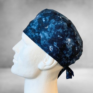 May include: A blue and white patterned surgical scrub hat with a galaxy print featuring stars, moons, and clouds. The hat has a tie at the back.