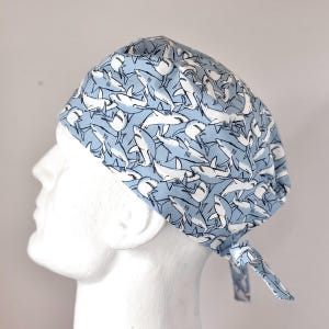 May include: A light blue surgical scrub hat with a white shark pattern. The hat has a tie at the back.