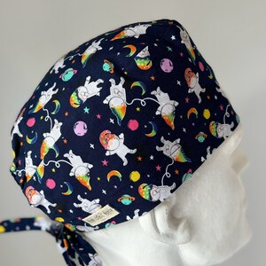May include: A navy blue surgical scrub cap featuring a whimsical space-themed pattern. The design includes astronauts with rainbow-colored trails, planets, stars, and crescent moons. The cap has a white lining and a tie closure.
