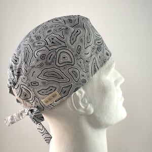 May include: A white and black patterned surgical scrub hat with a black and white tie. The hat has a label that says "The Scrub Hut".
